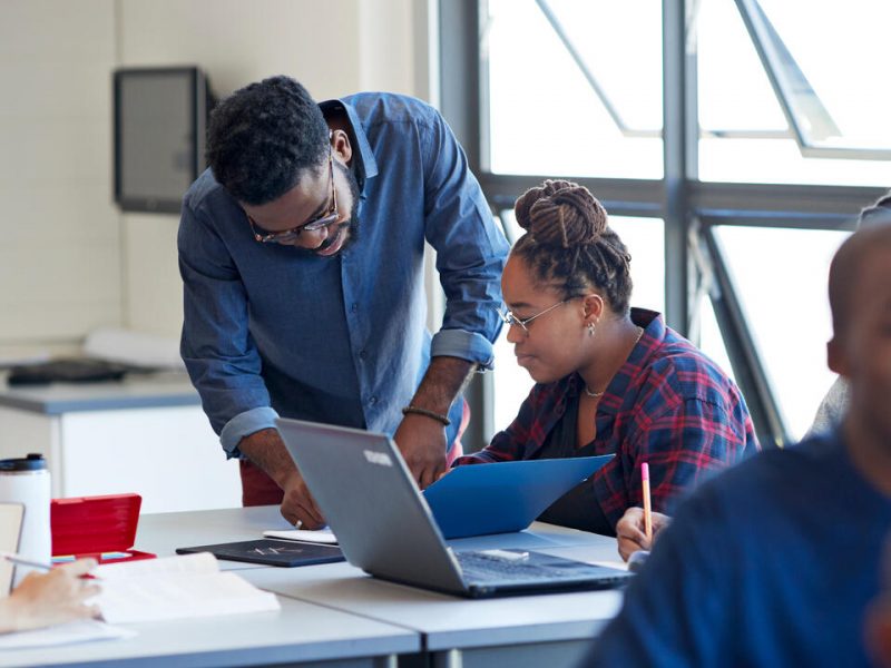 Young male teacher explaining female student at desk in university classroom